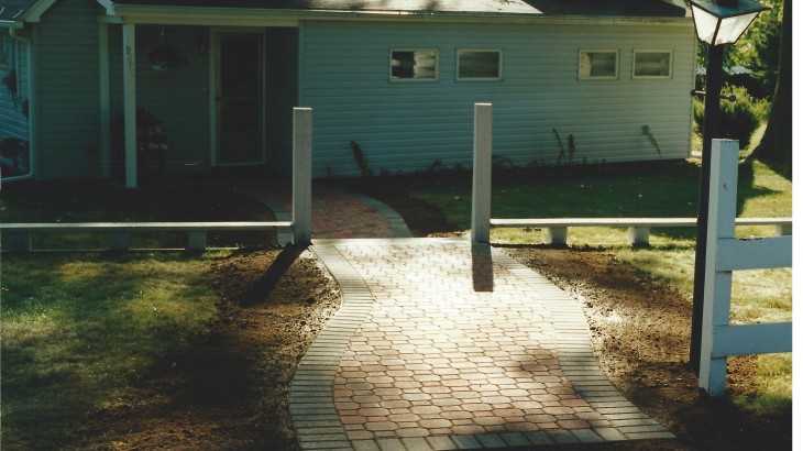 A brick pathway going thorugh a fence and leading up to a home
