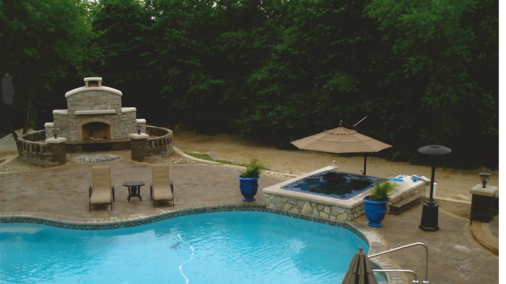 An inground pool surrounded by brick with a stone hot tup and brick fireplace