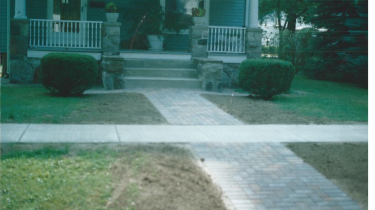 A brick paved walkway leading up to a house that has a concrete sidewalk intersecting the two paths