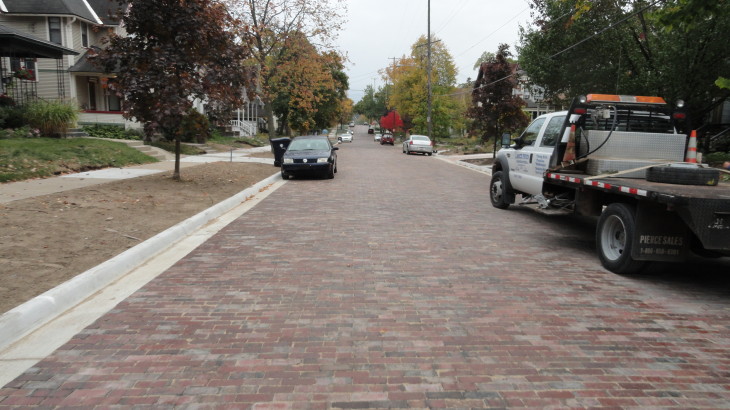 A finished brick road that runs through a subdivision with cars parked on the street outside of their houses