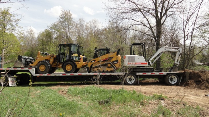 A series of loaders and excavators on a step deck stationed in a wooded area