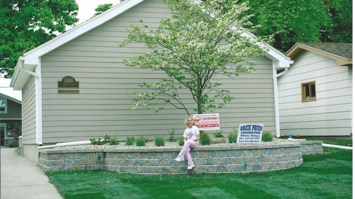 A girl sitting on a Retaining wall that houses trees and a sign that says Brick Paver Construction