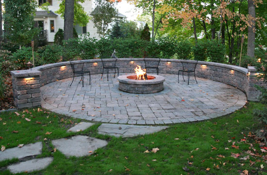 A stone path leading up to a circle of bricks with a circular fireplace in the middle with chairs sationed around the pit